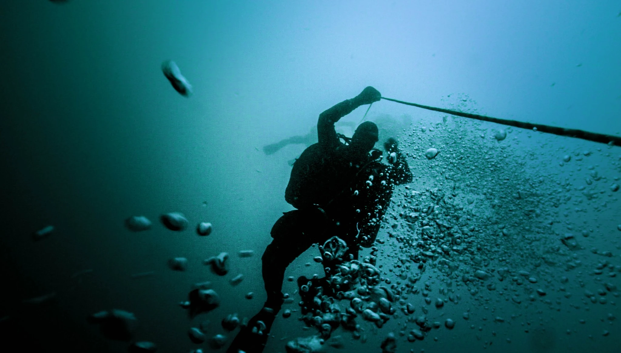Scuba diver holding an underwater rope while ascending through blue-green water, surrounded by rising air bubbles.